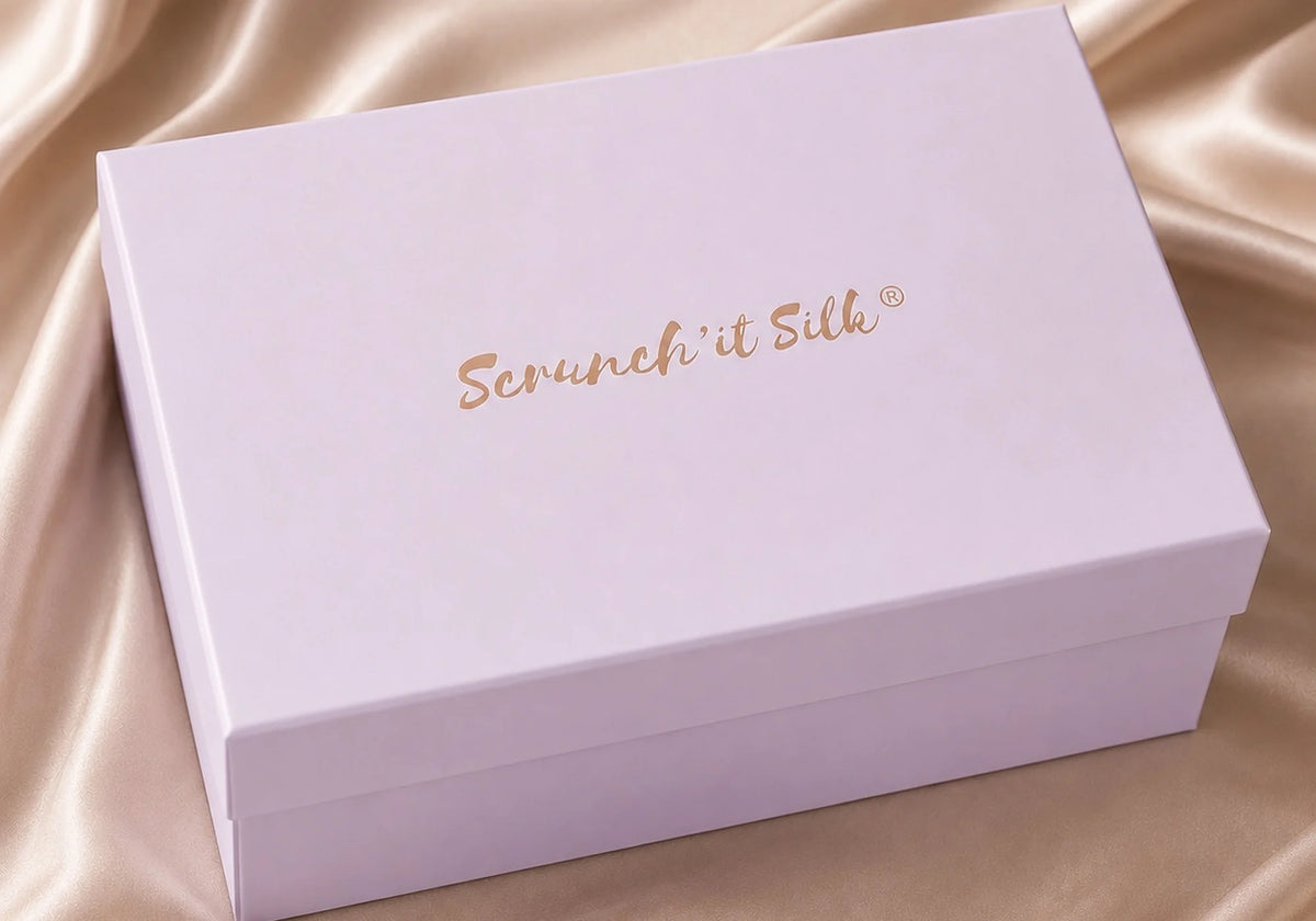 Boîte cadeau rose poudré Scrunch'it Silk avec logo bronze, posée sur un tissu en soie champagne.